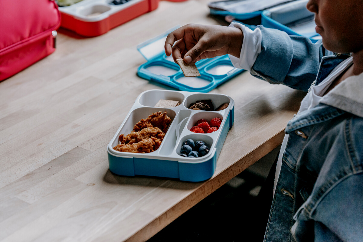 Crop black schoolchild with biscuit near lunch box